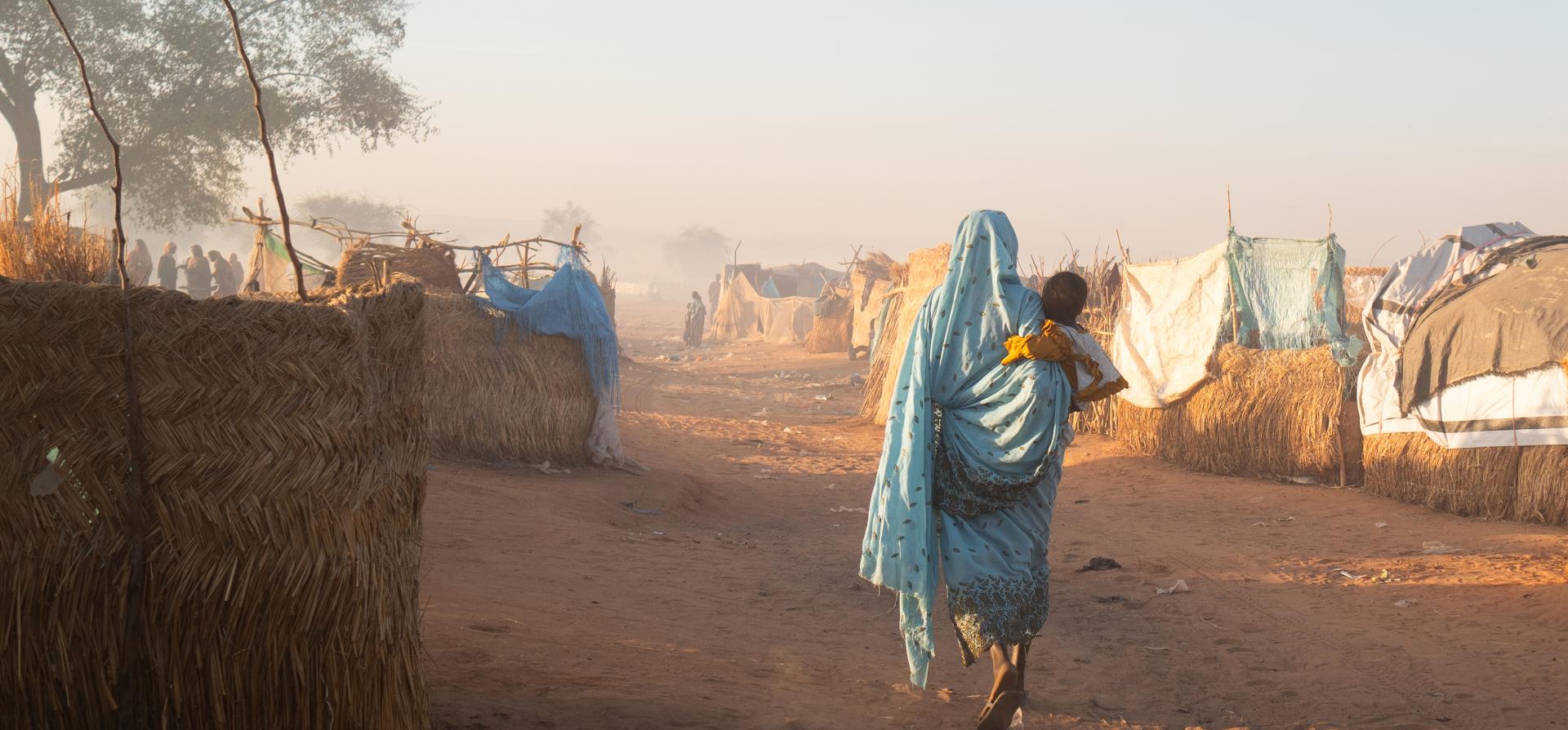 A woman walking through a refugee  camp with a baby on her back.